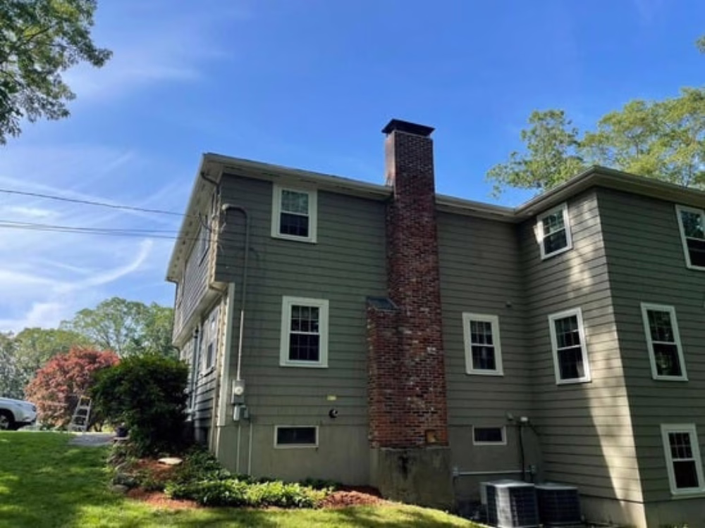 Side view of a two-story home in Holliston, MA after an exterior refresh, featuring a clean, even repaint in the original color with well-prepped siding and trim under a clear blue sky.